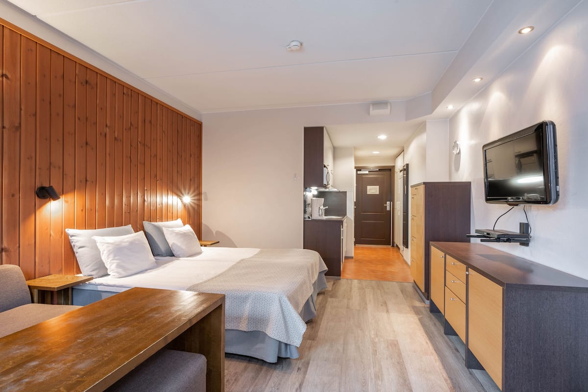 A hotel room with two beds, a wooden wall, a desk, a TV, and a kitchenette.