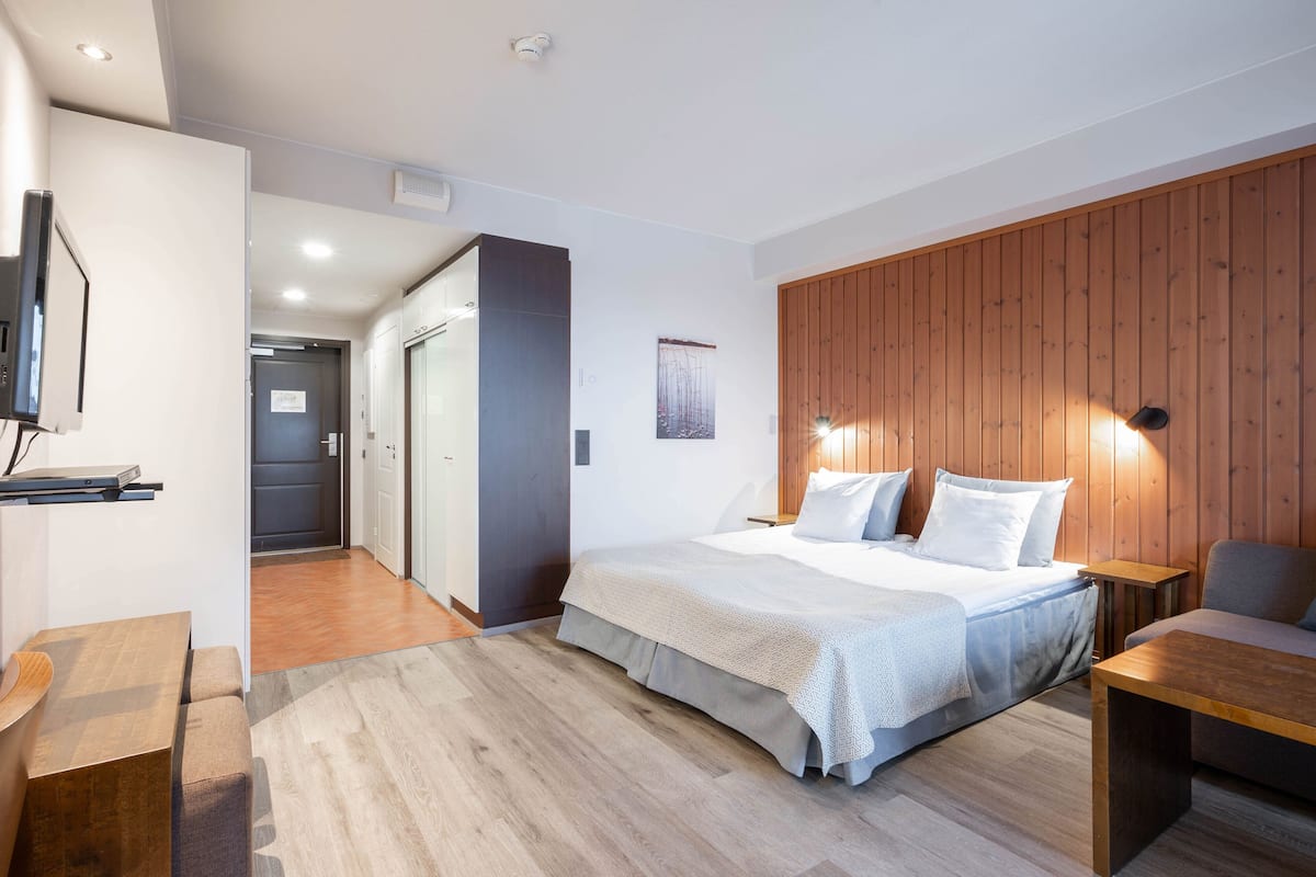 A modern hotel room with a large bed, a wooden accent wall, a flat-screen TV, and a small table.