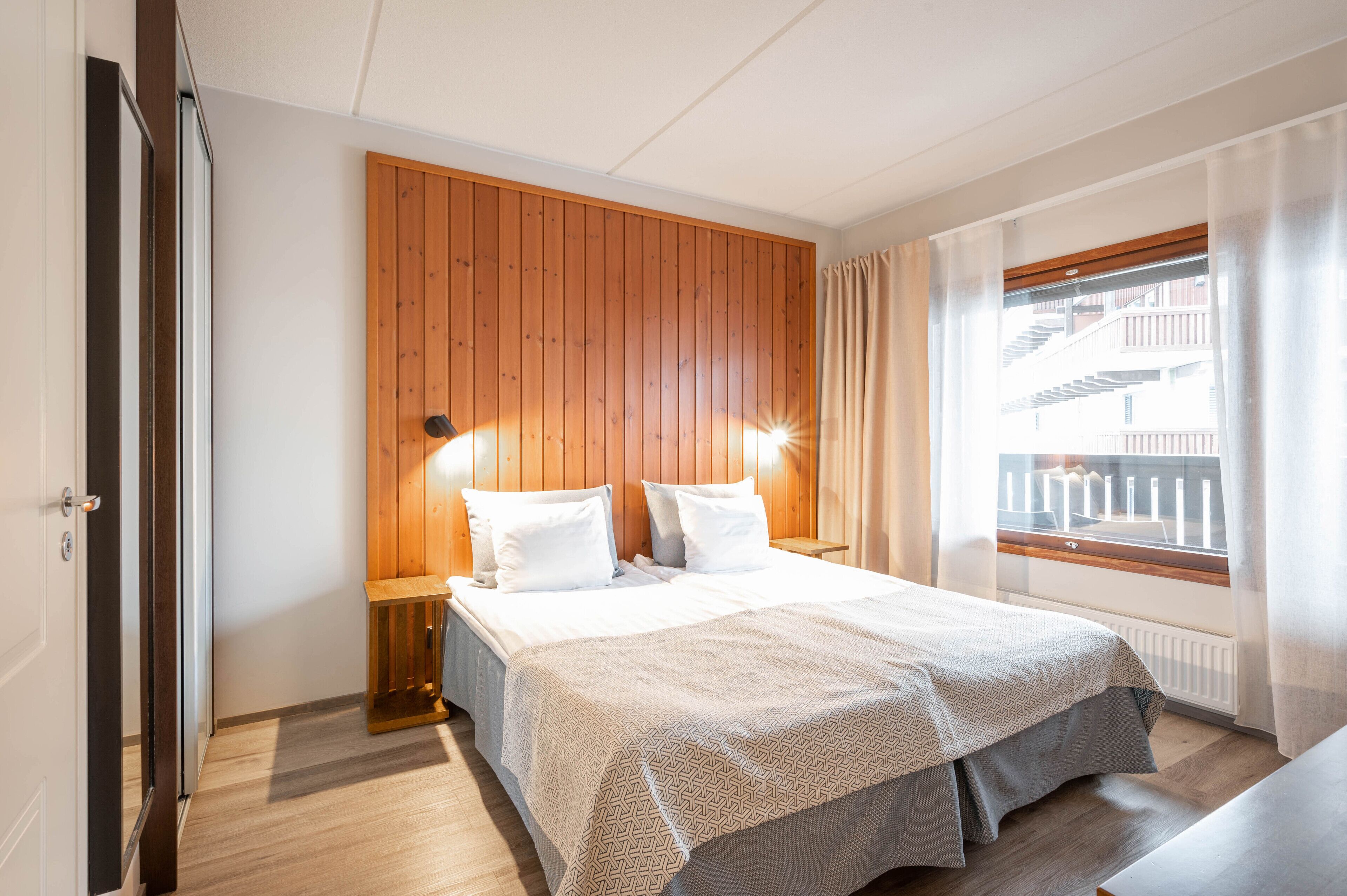 A hotel room with a wooden headboard, a large bed with white bedding, a nightstand with a lamp, a window with curtains, and a view of a building outside.