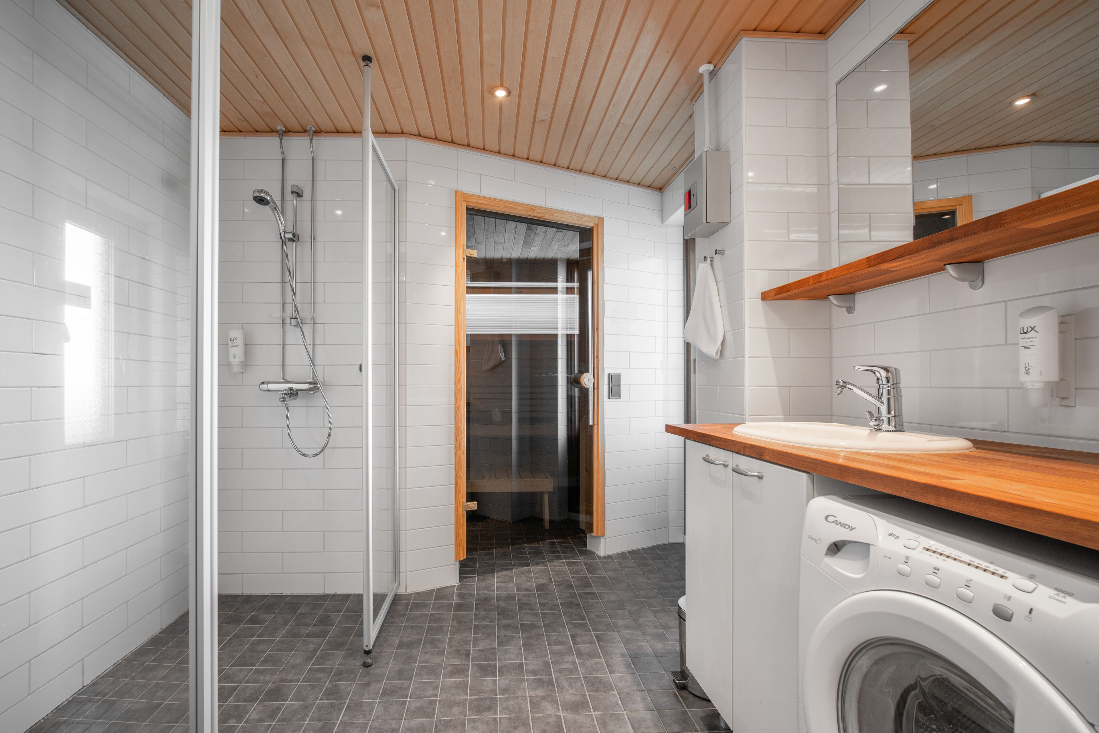 A modern bathroom with a glass shower enclosure, a wooden ceiling, a built-in washing machine, and a wooden countertop above the sink.