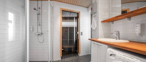 A modern bathroom with a glass shower enclosure, a wooden ceiling, a built-in washing machine, and a wooden countertop above the sink.