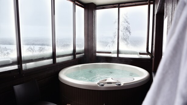 A hot tub in a corner with large windows offering a snowy landscape view. - Ski-Inn RukaVillage (Kuusamo)