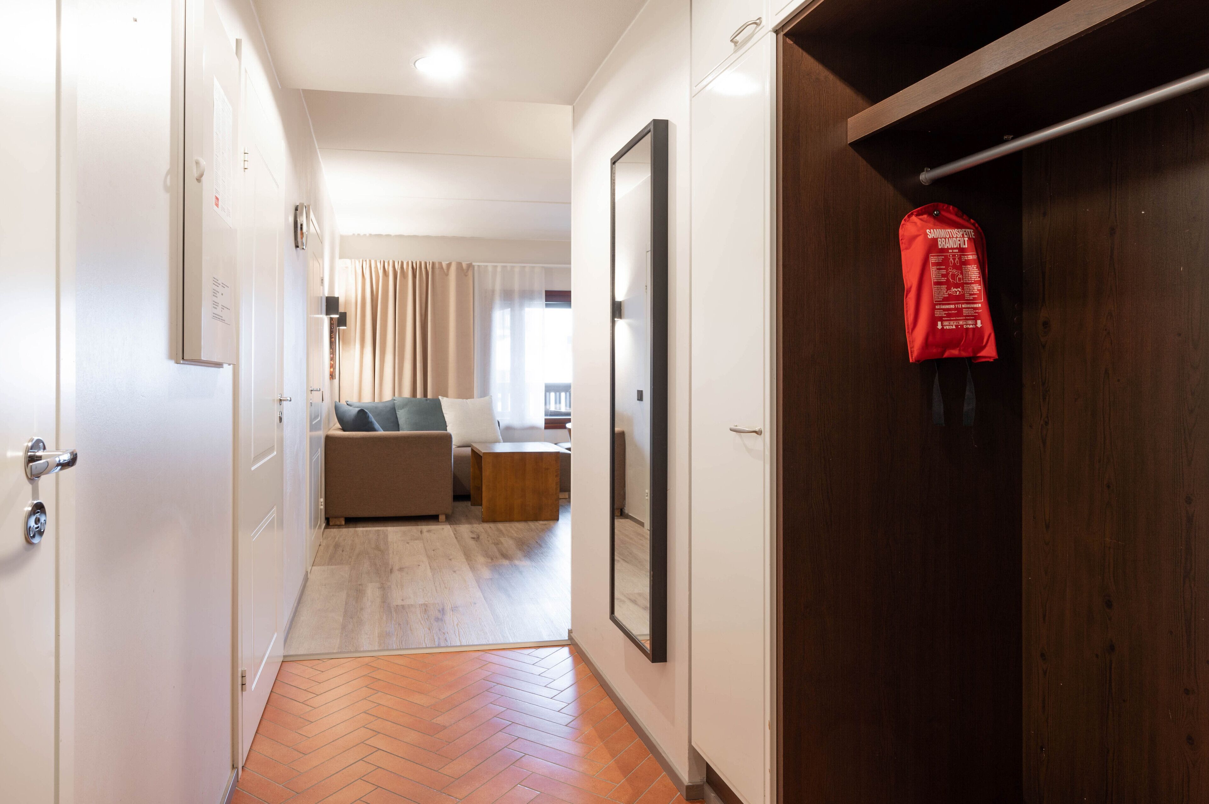 A hotel room with a wooden wardrobe, a beige sofa, a small wooden table, and a red bag hanging on the wardrobe.