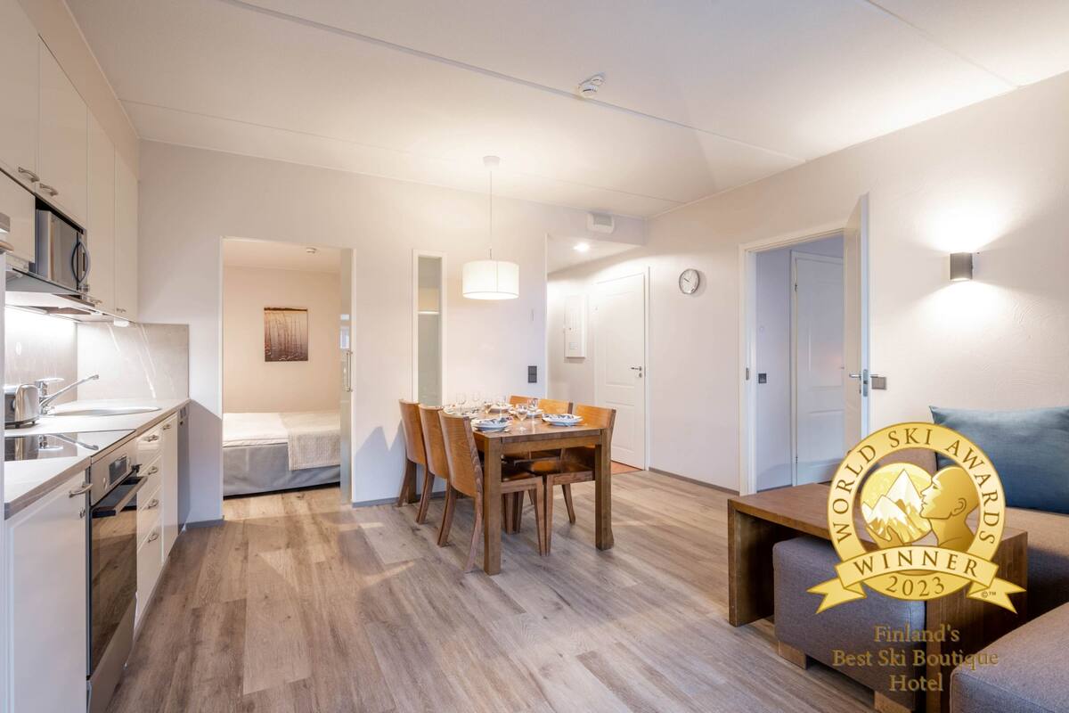 A modern hotel room with a kitchenette, a dining area with a wooden table and chairs, a sofa, and a bed.