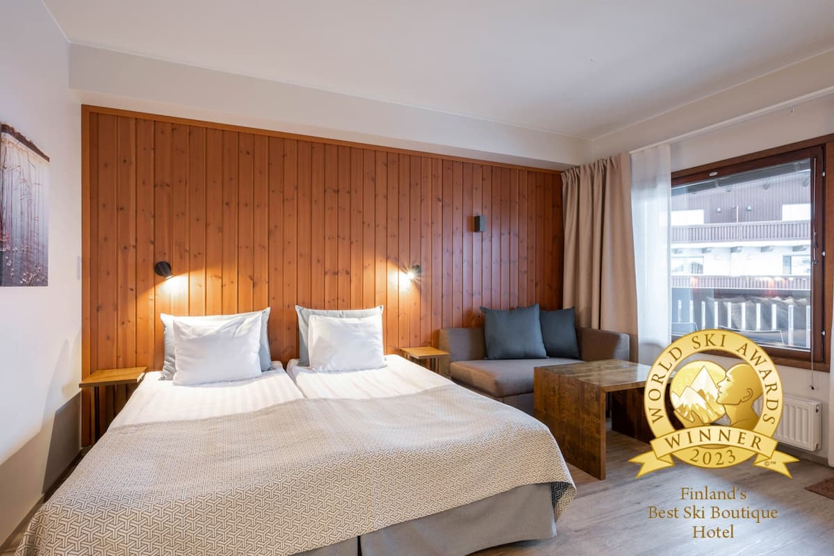 A hotel room with a wooden panel wall, a large bed with two pillows, a sofa, a small table, and a window with curtains.