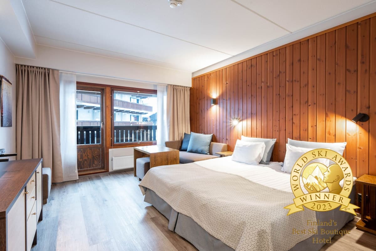 A hotel room with a wooden paneled wall, a large bed with a patterned bedspread, a sofa, a small table, and a view of a balcony with a railing.