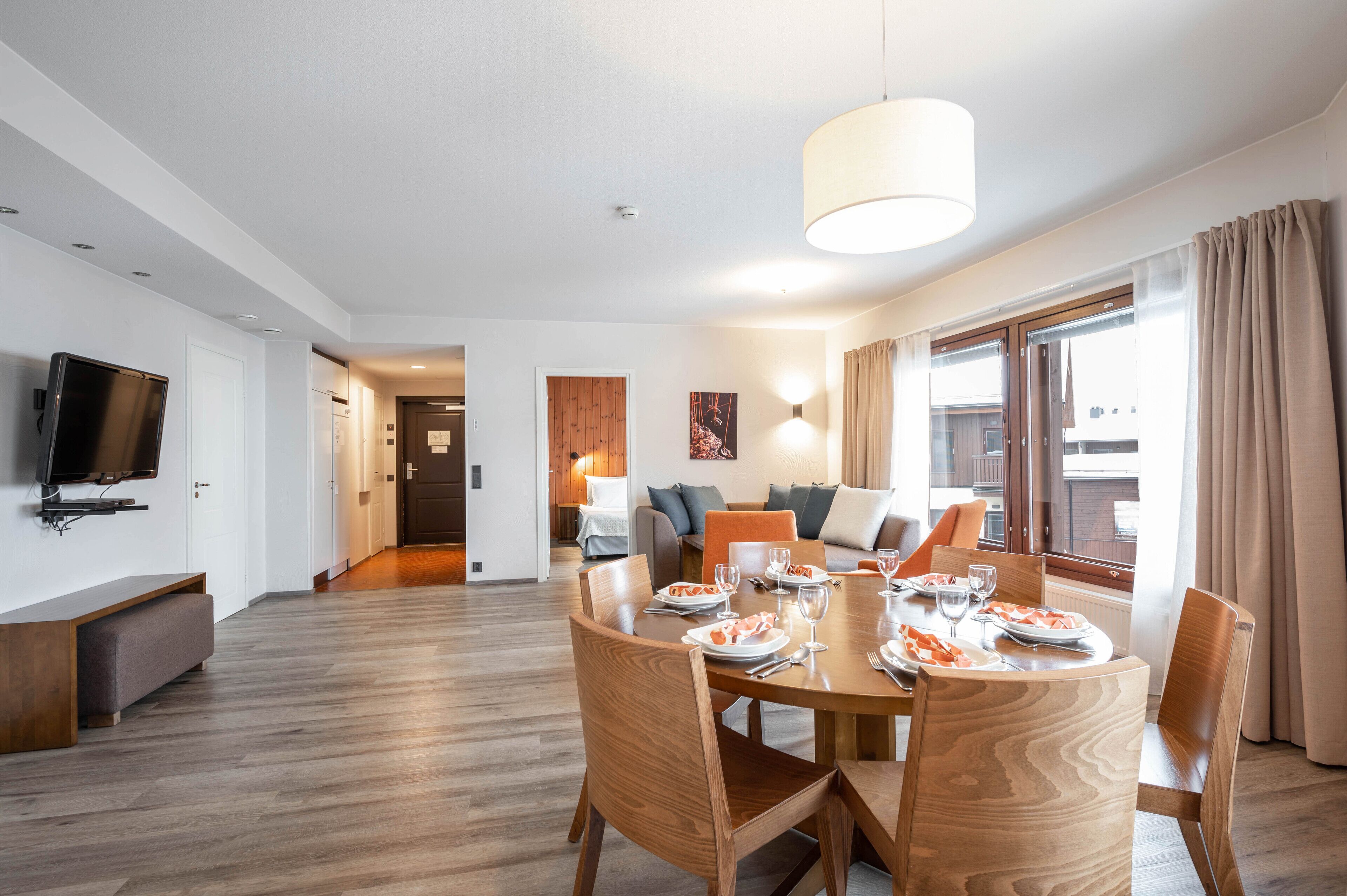 Un espace repas moderne avec une table en bois dressée pour quatre personnes, un téléviseur à écran plat fixé au mur et un canapé en arrière-plan.