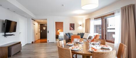Un espace repas moderne avec une table en bois dressée pour quatre personnes, un téléviseur à écran plat fixé au mur et un canapé en arrière-plan.