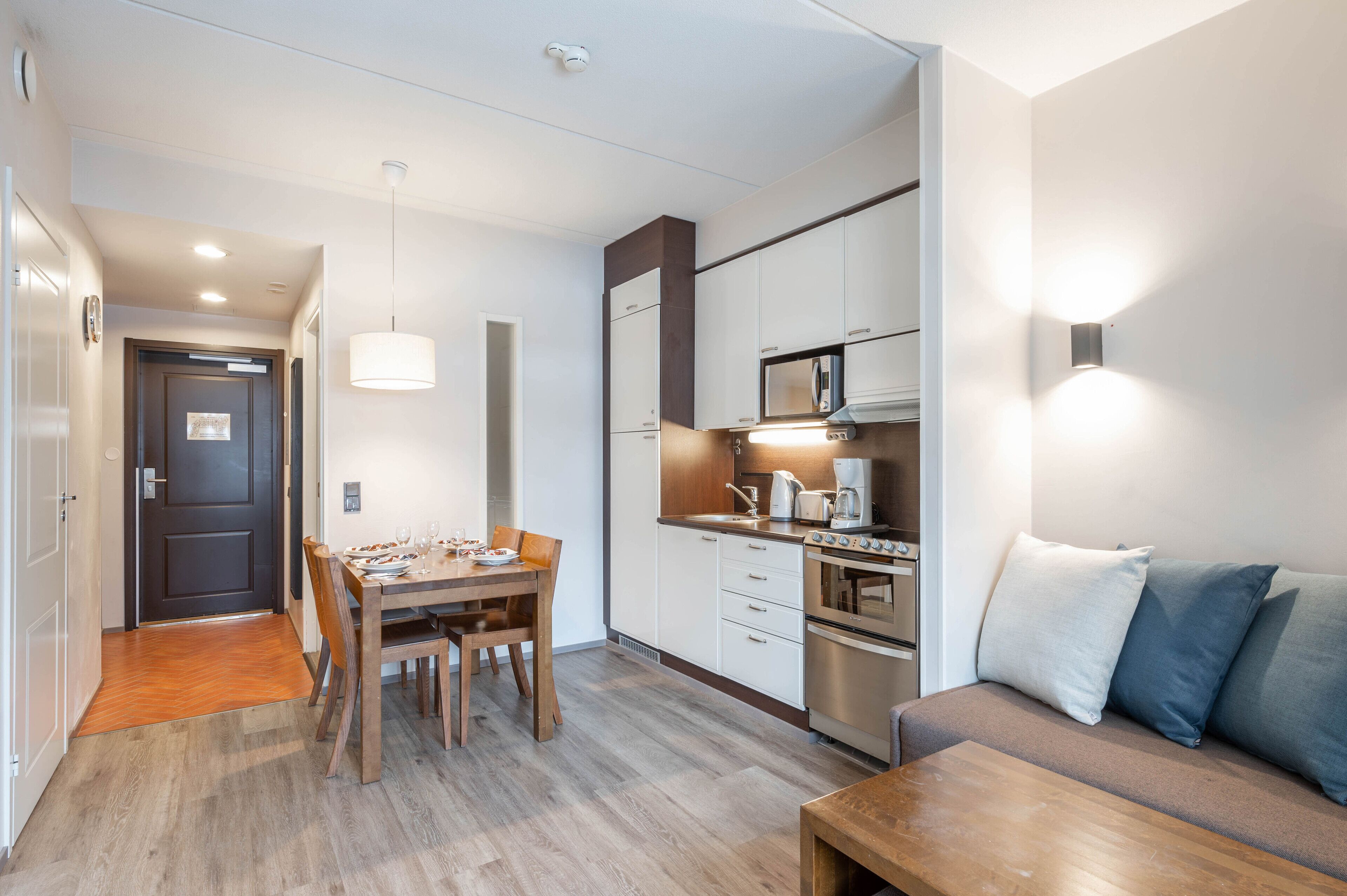 A compact kitchen with modern appliances, a dining area with a wooden table and chairs, and a sofa with cushions.