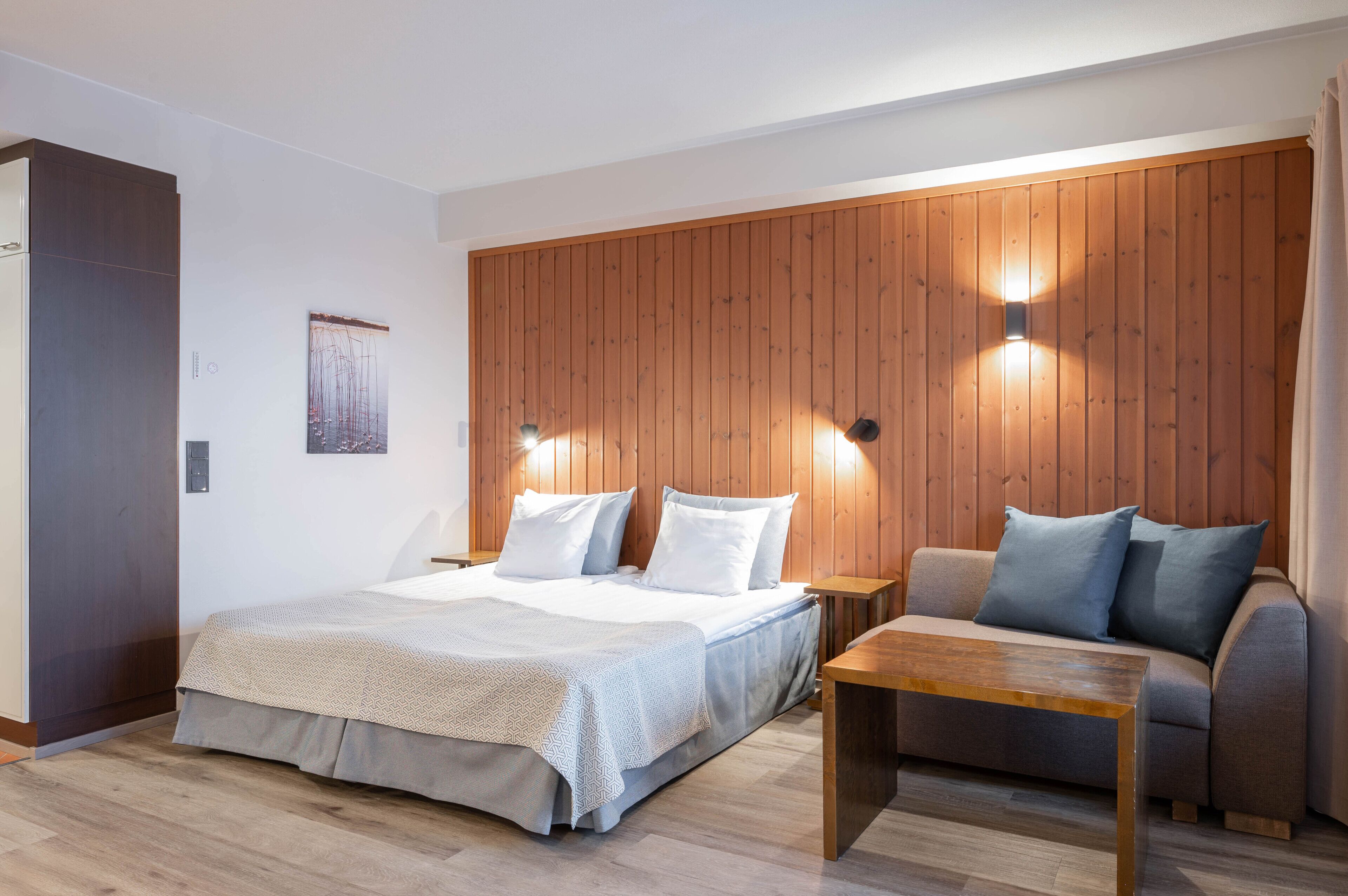 Een moderne slaapkamer met een wand van houten panelen, een bed met witte beddengoed, een grijze bank met blauwe kussens, en een houten salontafel.