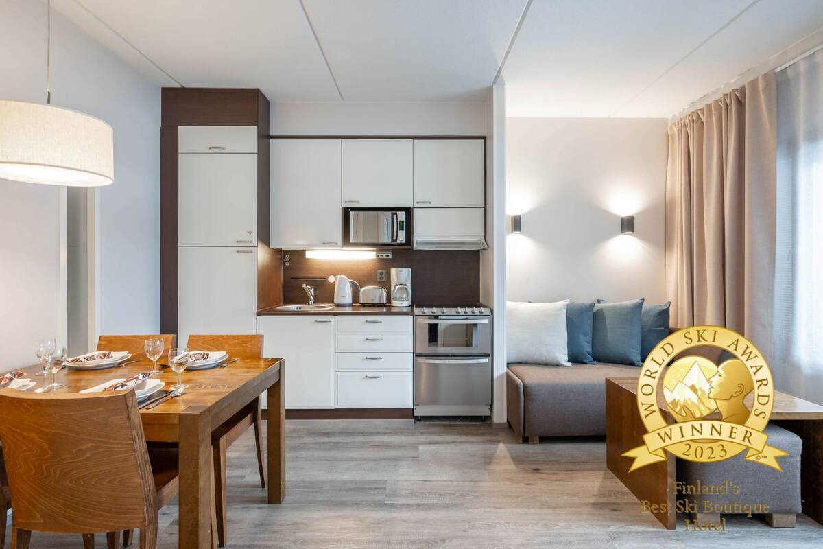 A modern hotel room with a dining area, kitchenette, and a sofa. It features a kitchen with white cabinets, a stove, and a microwave. There is a dining table set with plates and glasses. The room has a neutral color scheme with beige curtains and a light-colored sofa.