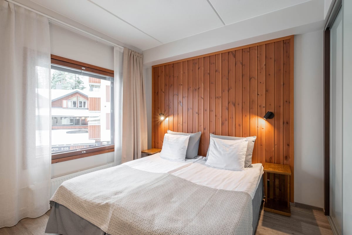 A bedroom with a wooden headboard, two bedside tables, a bed with patterned bedding, a window with curtains, and a view of buildings outside.
