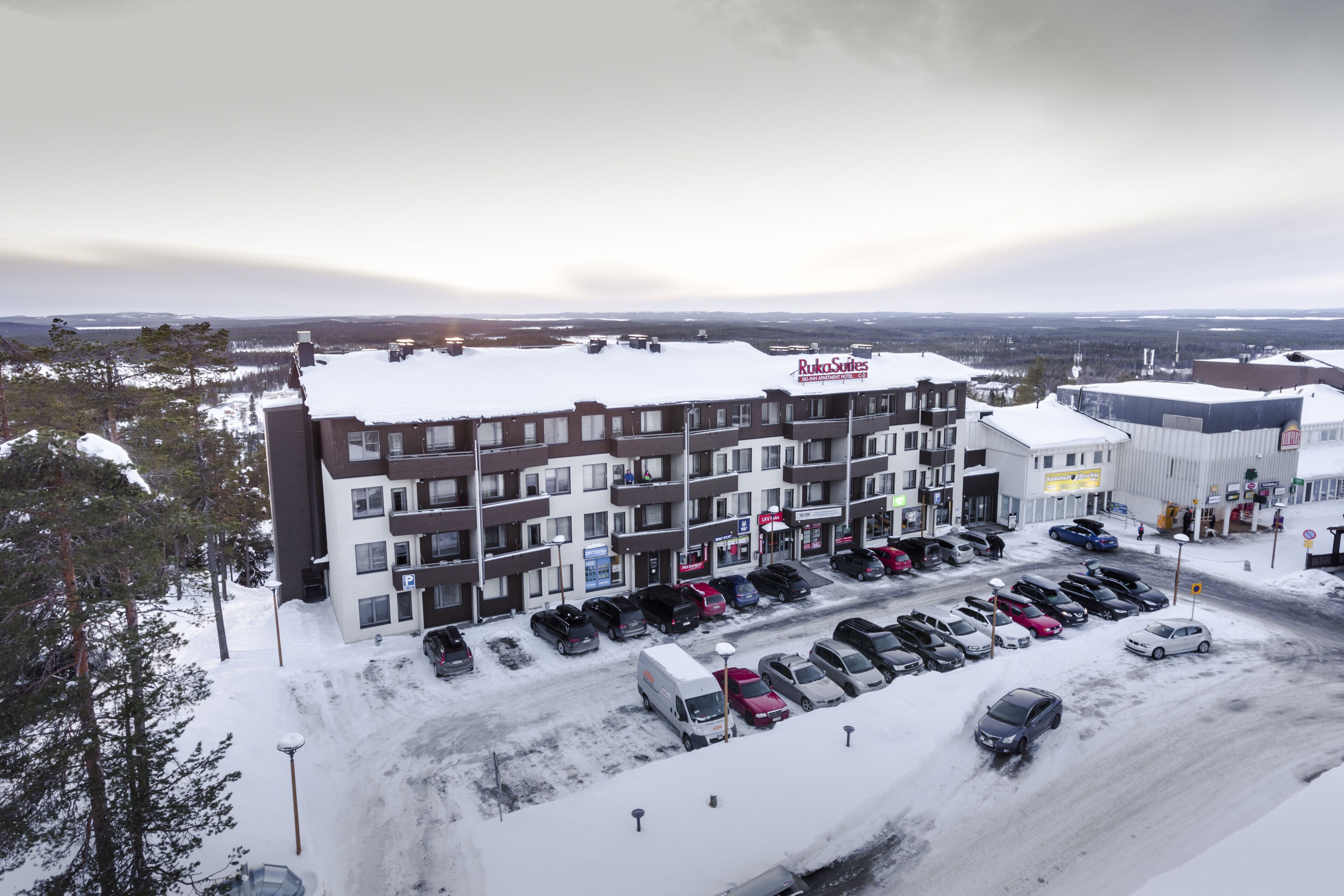 Un hotel a più piani con un parcheggio pieno di auto, circondato da alberi e edifici innevati.