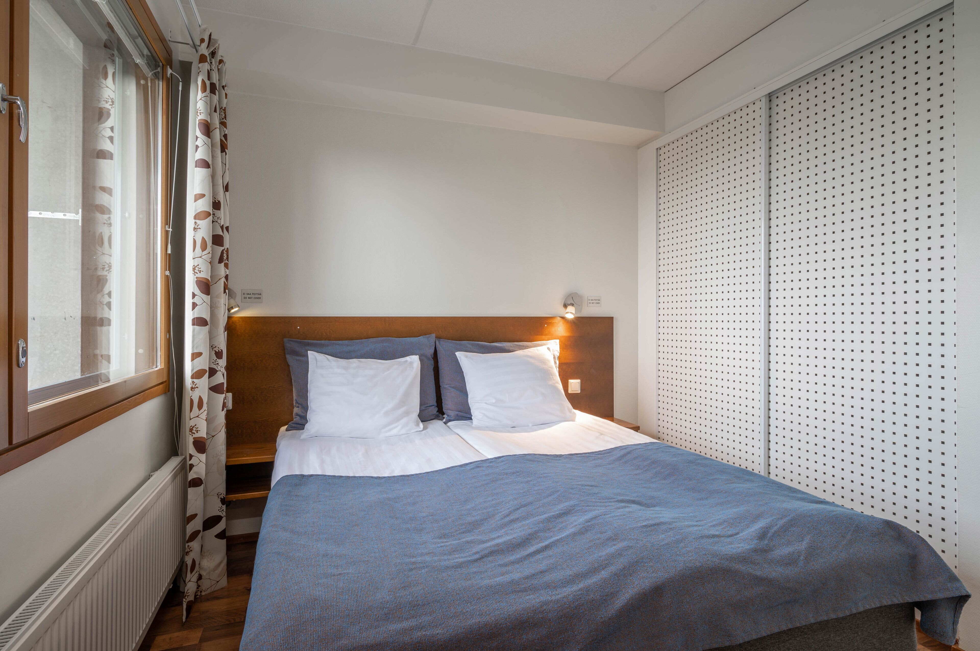 A neatly made bed with a wooden headboard, a window with curtains, and a wall with a perforated design.