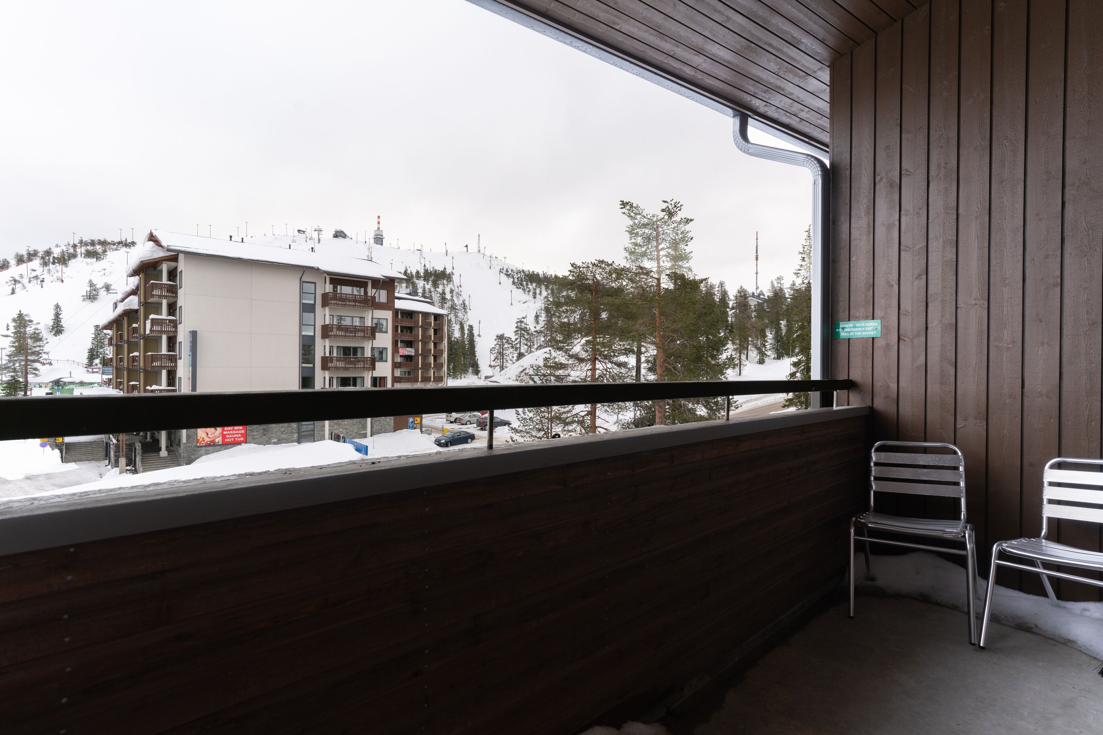 Ein Balkon mit Blick auf einen verschneiten Berg und ein Gebäude. Auf dem Balkon befinden sich zwei Metallstühle.