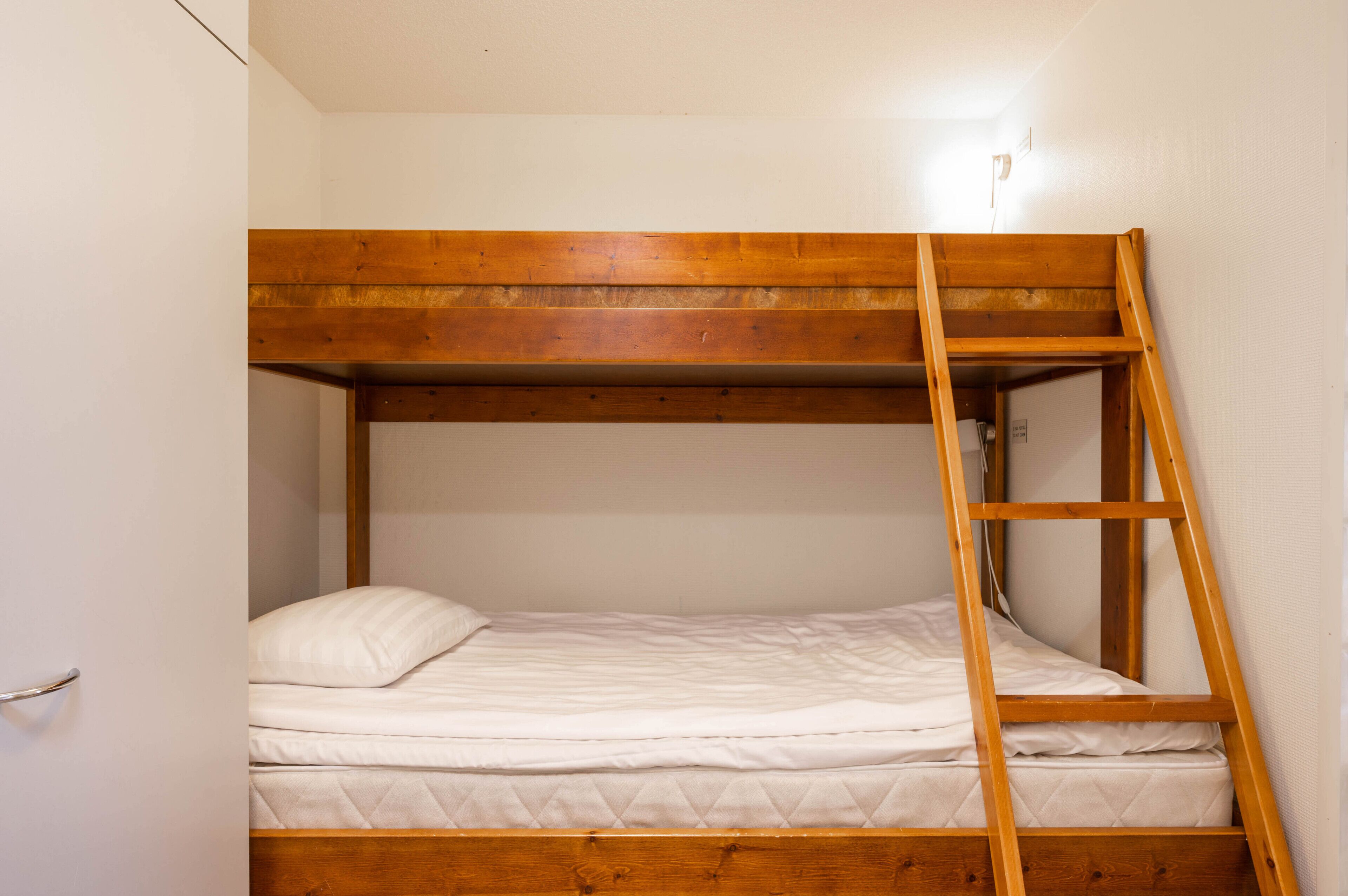 Een stapelbed met een houten trap, wit beddengoed en een witte muur.