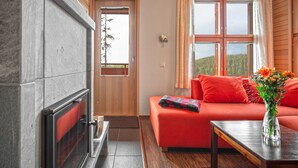 A cozy room with a fireplace, a sofa, a wooden table, and a window with a view of trees. - Ski-Inn AurinkoRinne (Kuusamo)
