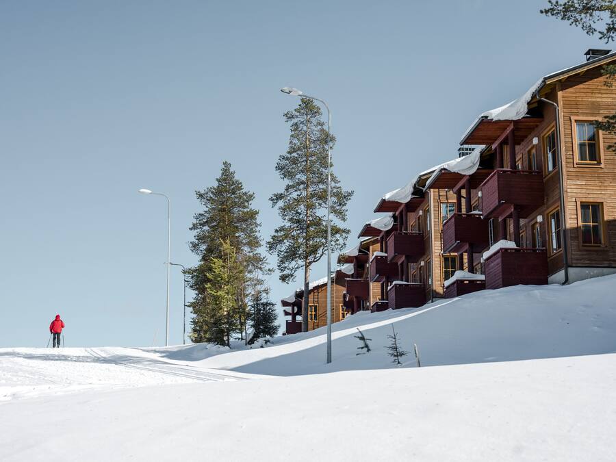 A snow-covered slope with a person skiing, wooden buildings with balconies, and tall pine trees.