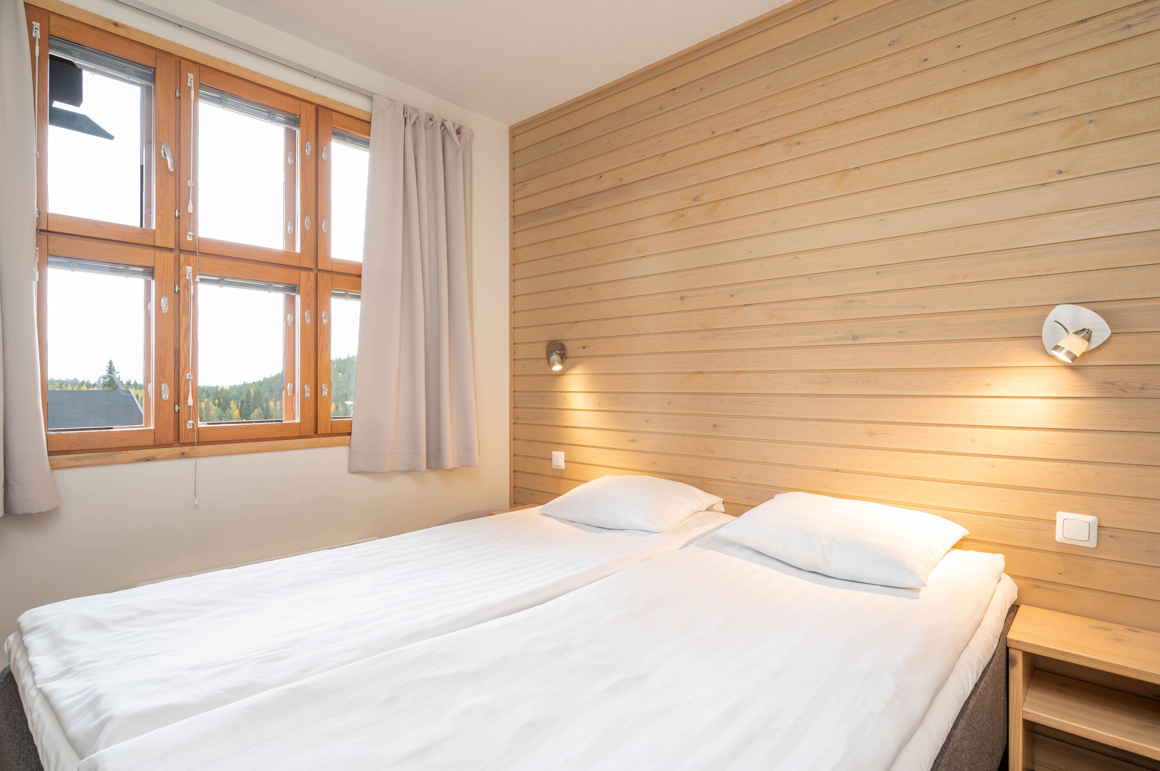 Un lit double avec du linge de lit blanc, une tête de lit en bois, une table de chevet, une lampe fixée au mur et une fenêtre avec des rideaux.
