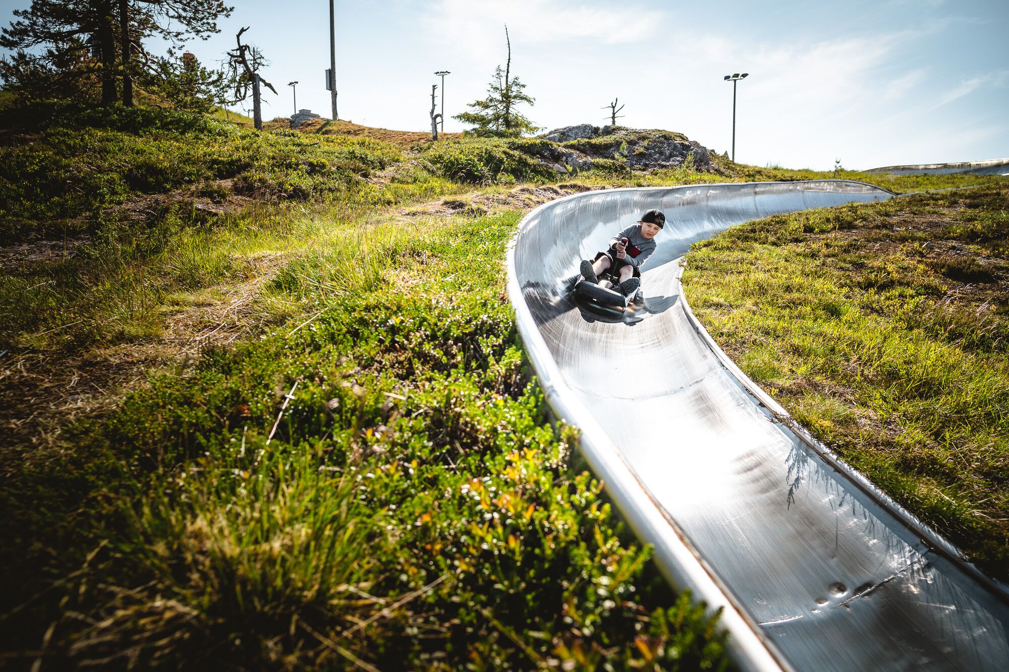 A person sliding down a metal slide in a natural setting with grass and trees.
