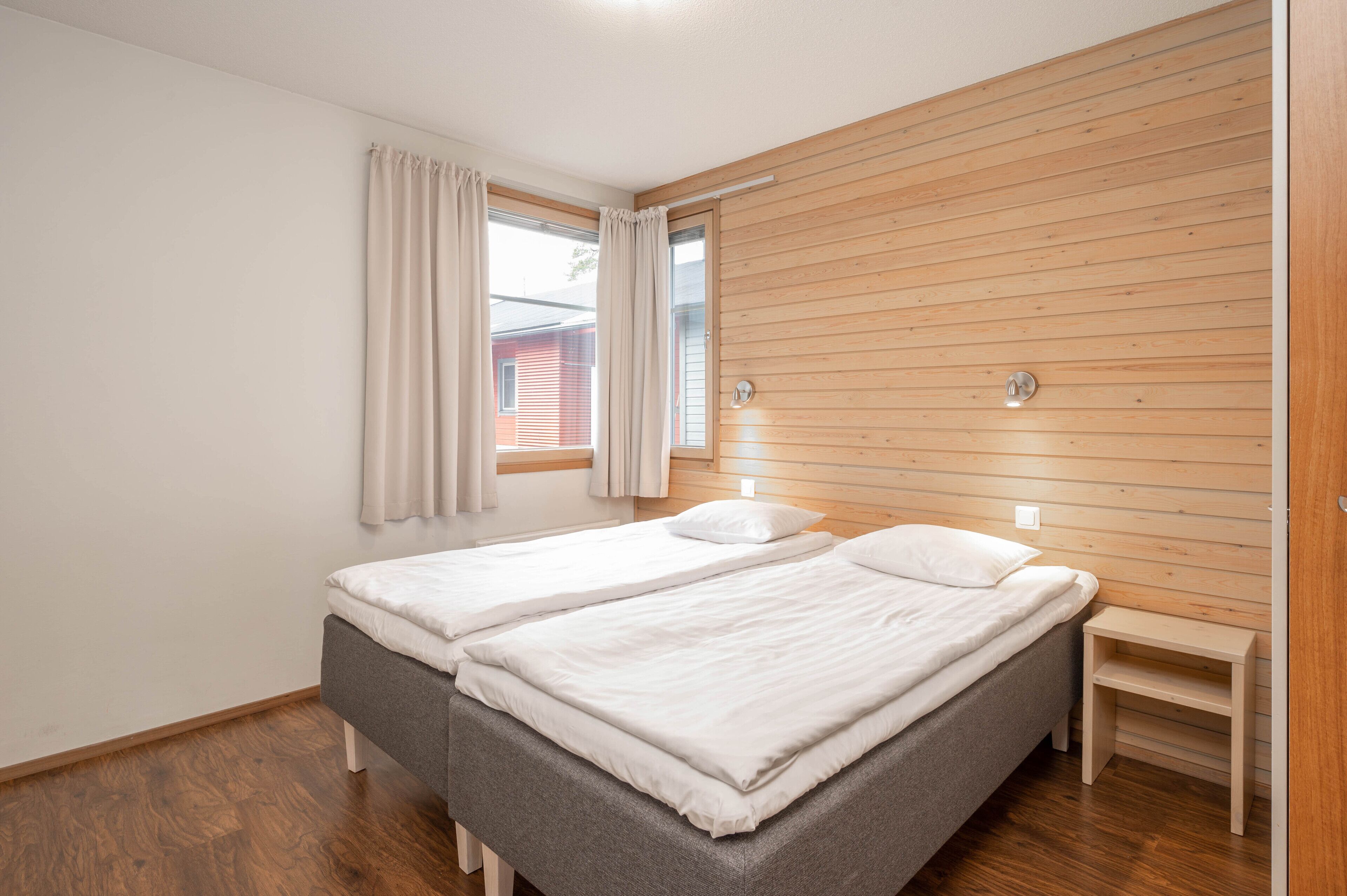 A bedroom with two single beds, wooden paneling on the walls, a window with curtains, and a small wooden nightstand.