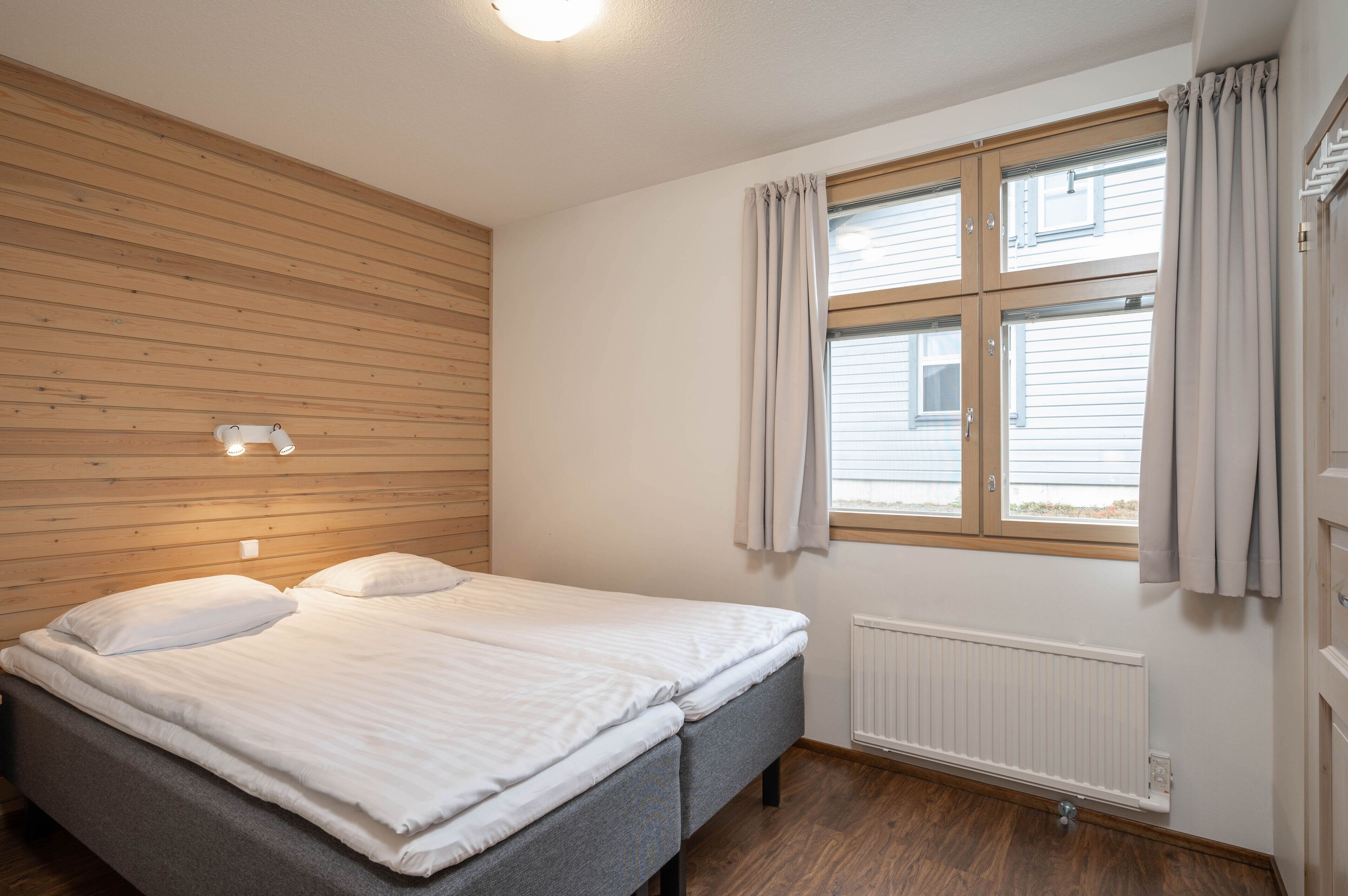 Et soveværelse med en seng, et vindue med gardiner, en trævæg og en lampe.