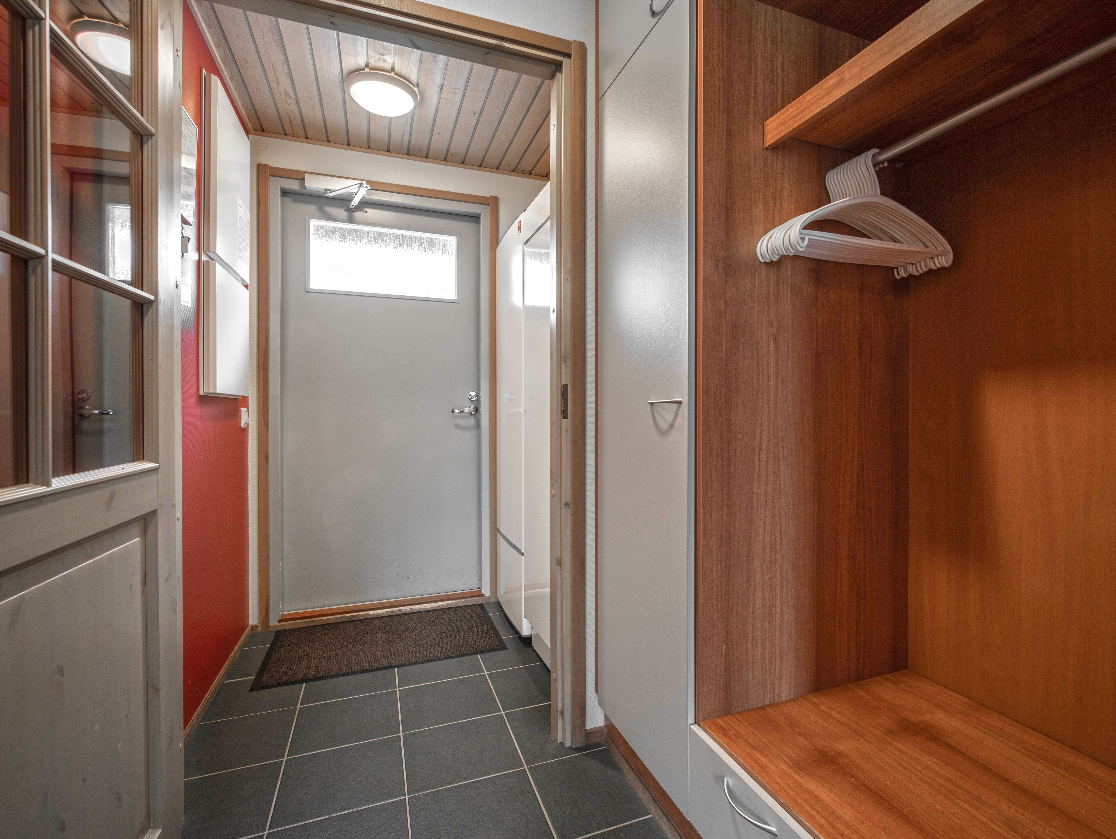 Een gang met een houten vloer, een witte deur met een raam, en een houten garderobe met kledingroede.