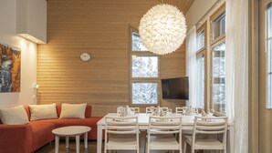 A modern living room with a wooden ceiling, a sofa, a dining table with chairs, and a large window with a view of snow-covered trees. - Ski-Inn MastonAitio (Kuusamo)