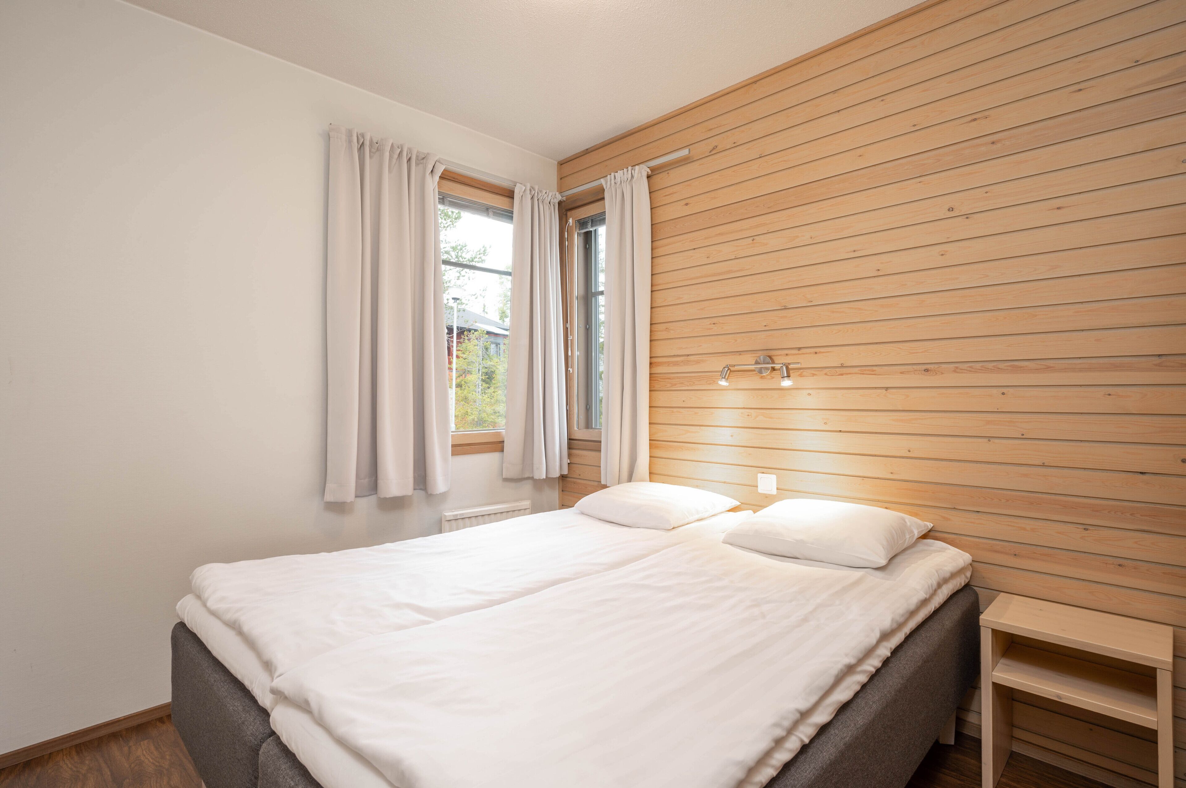 Un dormitorio con cama, pared de paneles de madera, ventana con cortinas y una mesita de noche.