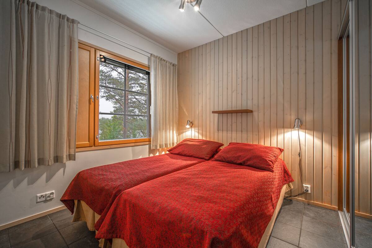 A room with two beds, a window with curtains, a wall-mounted shelf, and a wall-mounted light.