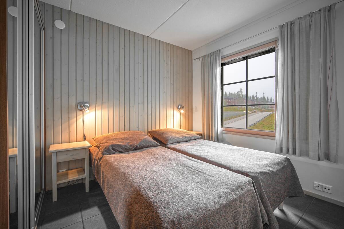 A double bed with a patterned bedspread, two bedside tables, a window with curtains, and a view of a road and trees outside.