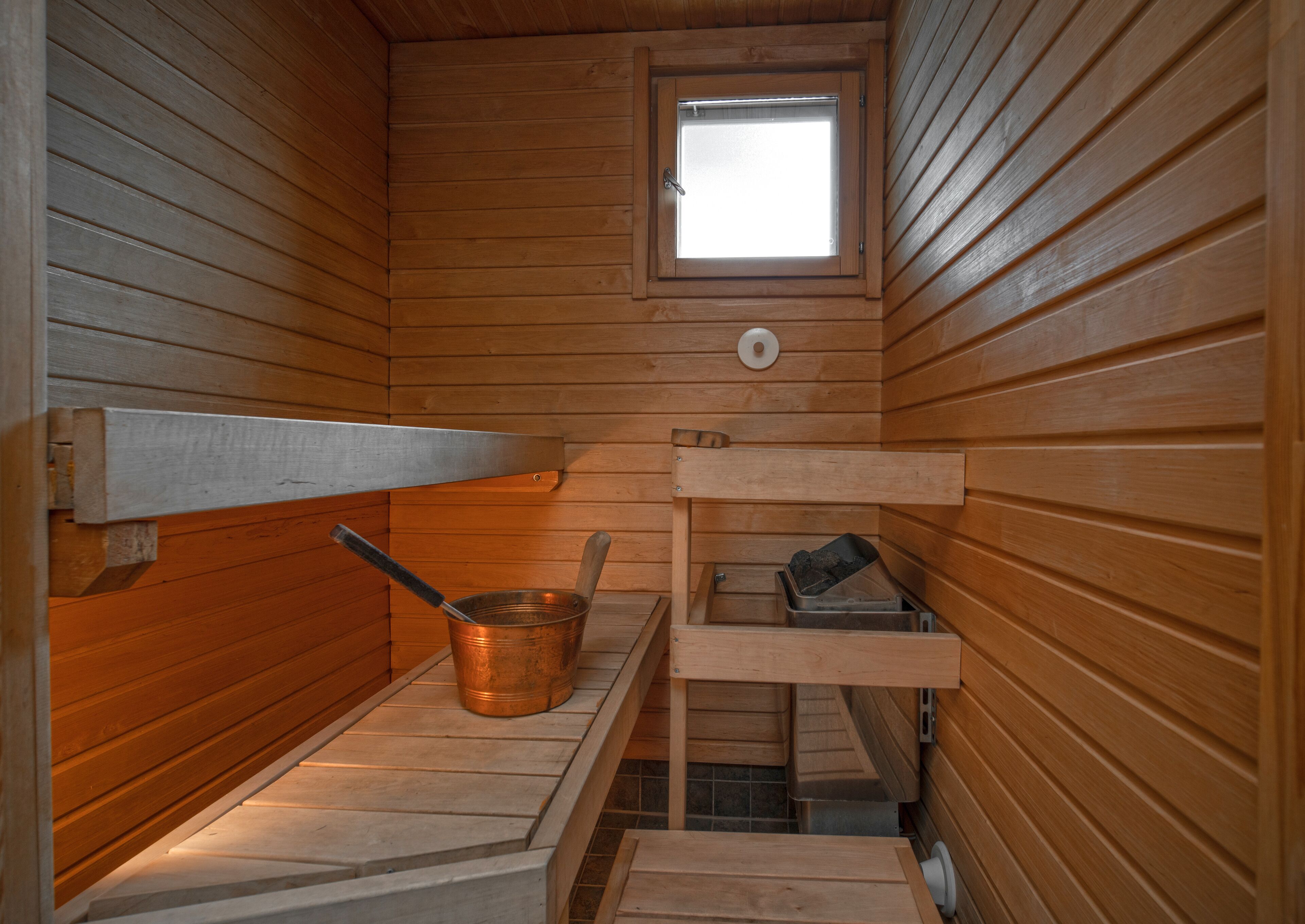 Una sauna de madera con ventana, un balde de metal y un estante de madera.