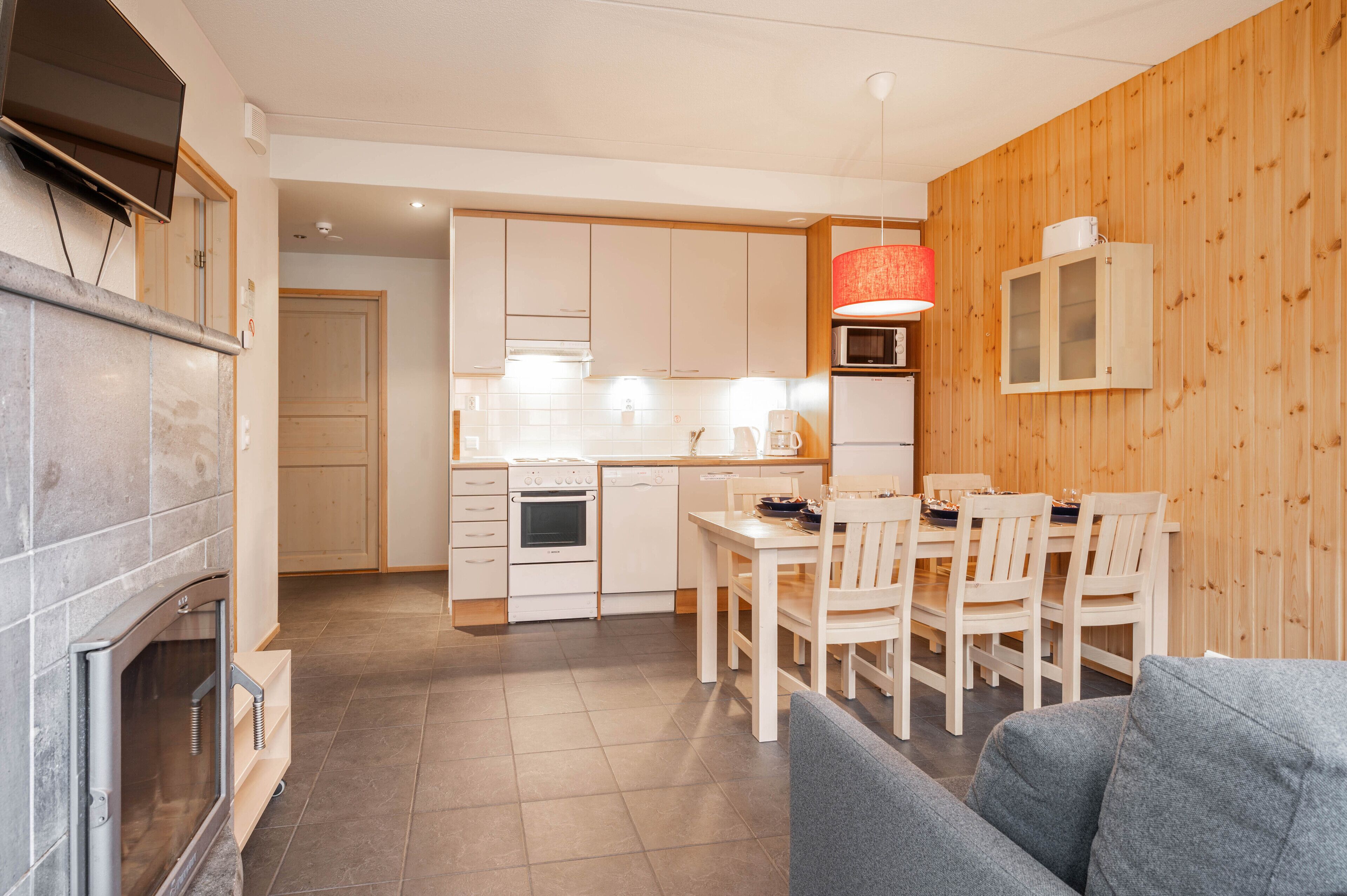 A modern kitchen with white cabinets, a dining area with a wooden table and chairs, and a grey sofa in the living room.