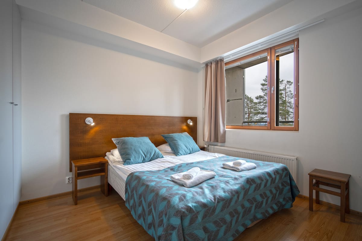 Un dormitorio con cabecera de madera, dos mesitas de noche, una cama con colcha estampada y una ventana con cortinas.