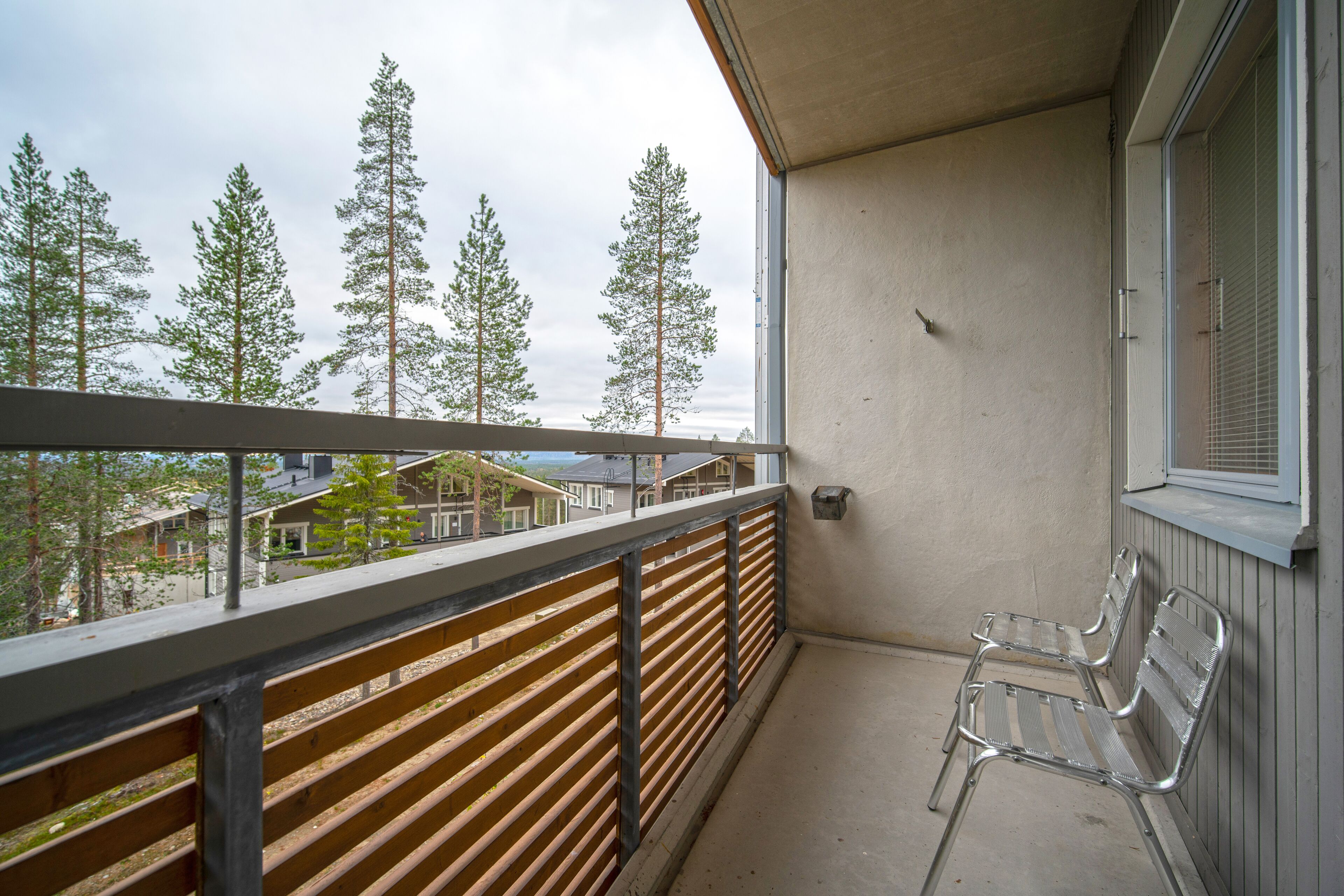 A balcony with a wooden railing, a metal bench, and a view of trees and houses.