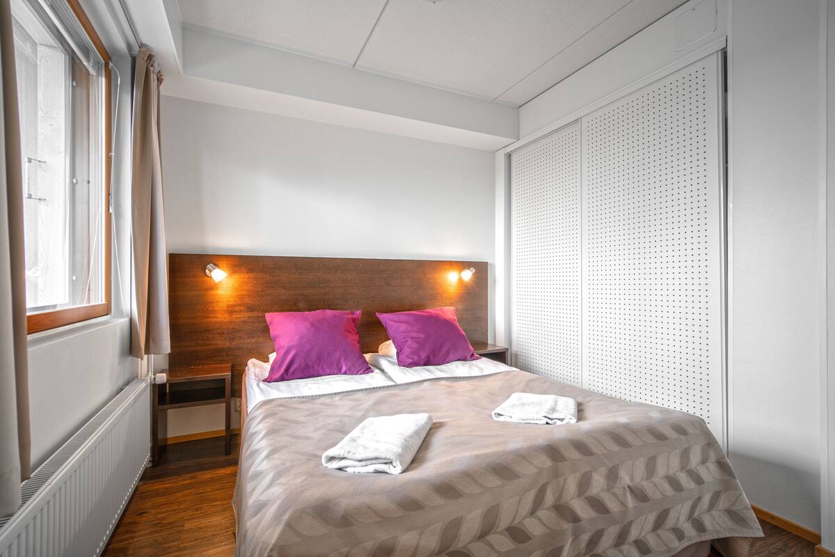 Habitación de hotel con cama, dos almohadas, mesita de noche, ventana y pared con panel perforado.