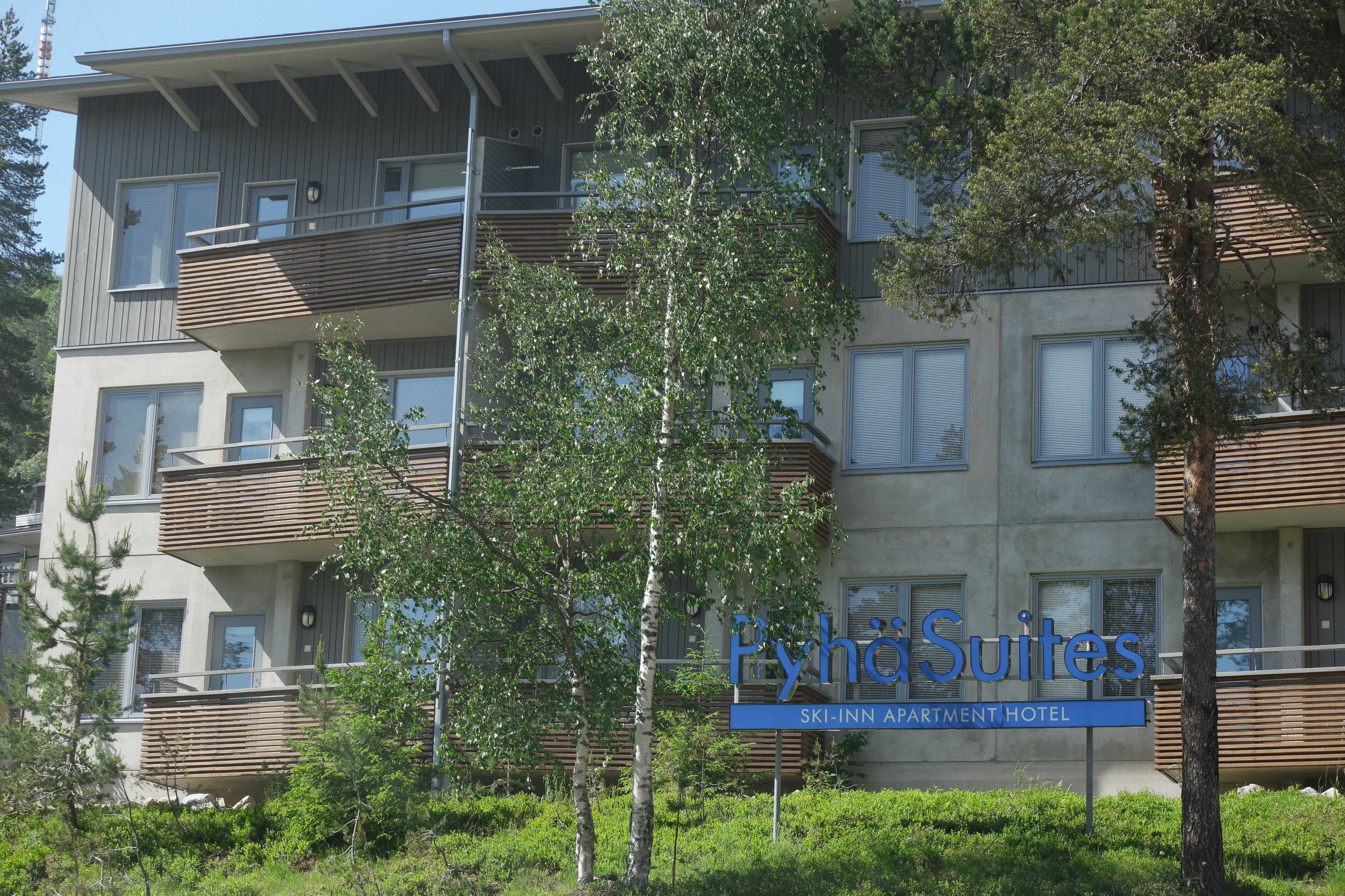 A multi-story hotel with balconies, a sign reading "PyhosSuites Ski-Inn Apartment Hotel," and surrounding greenery.