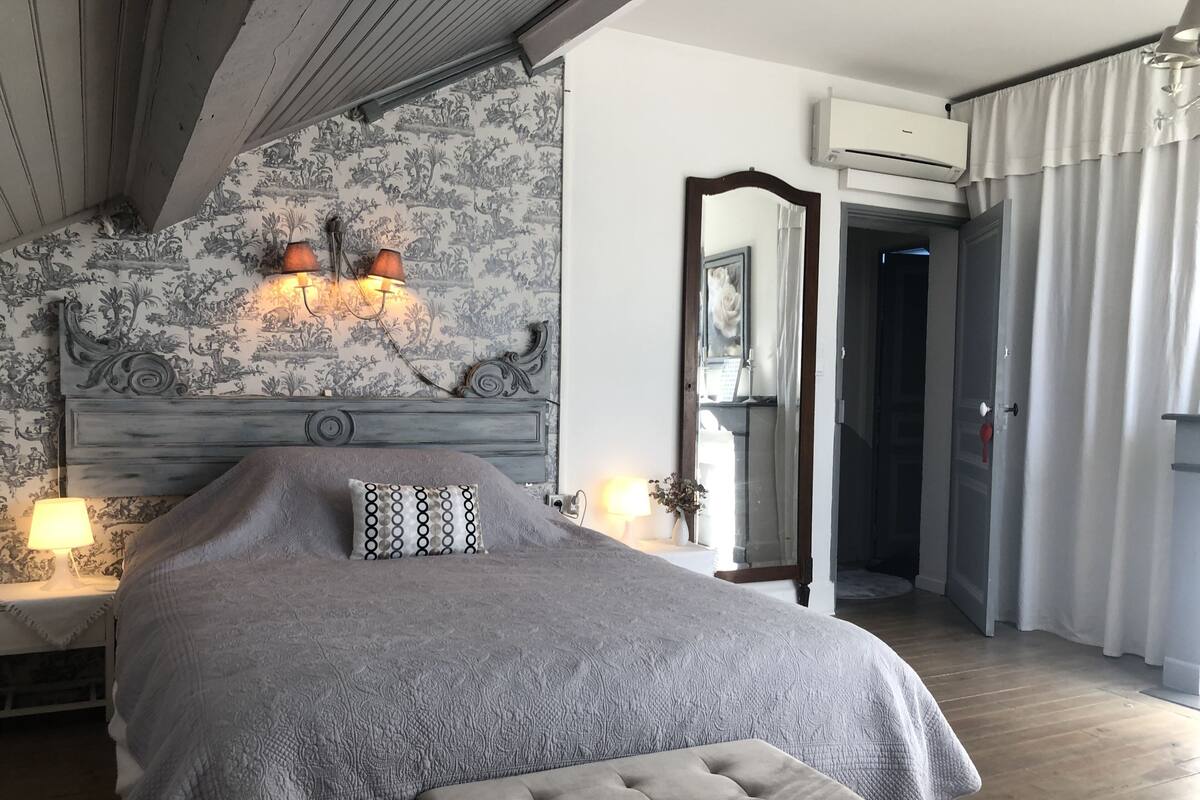 Une chambre à coucher avec un grand lit, une tête de lit agrémentée d’œuvres d’art murales, deux lampes de chevet, un miroir et un appareil de climatisation.