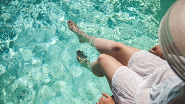 A person sitting by a pool with clear water, wearing a white shirt and shorts. - Au Bien Venu (Noe)