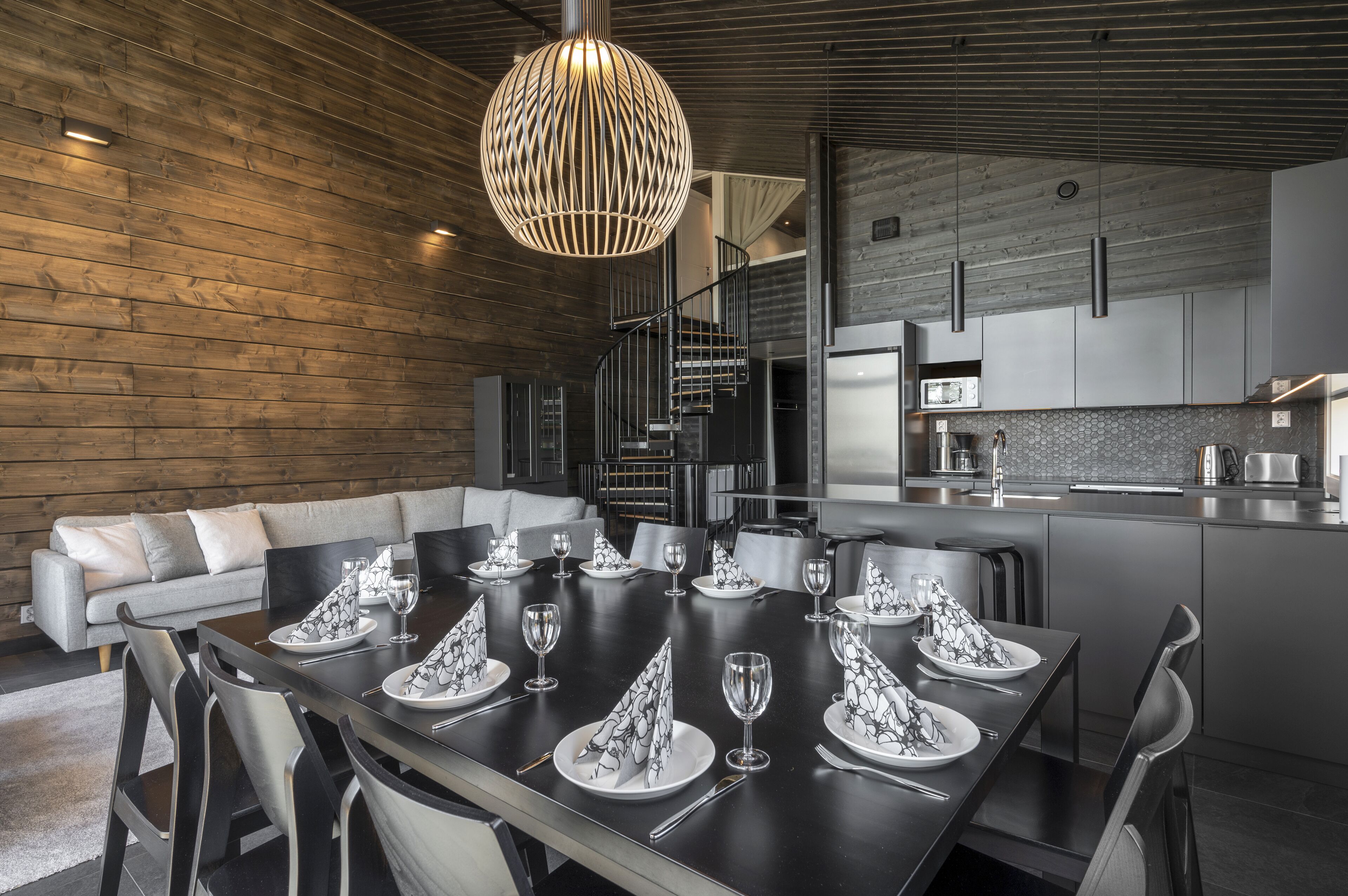 A modern dining area with a dark wooden table set for a meal, a staircase leading to an upper level, and a kitchen in the background.
