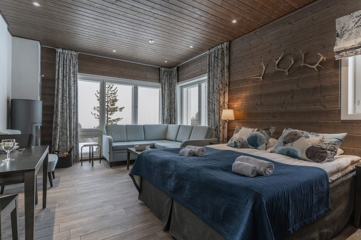 A modern bedroom with a wooden ceiling, a large bed with a blue bedspread, a grey sofa, and a dining area with a table and chairs.