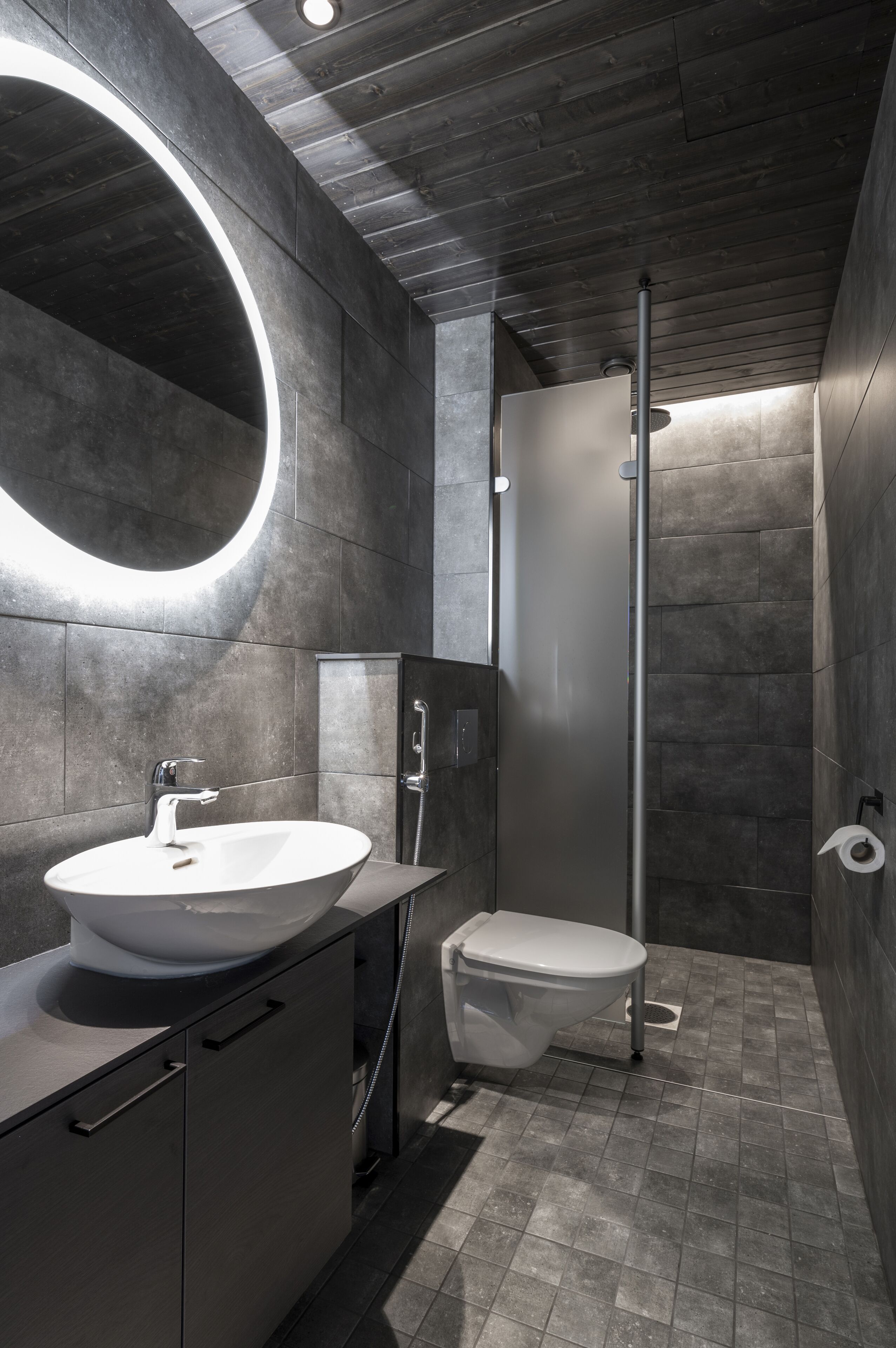 A modern bathroom with a large round mirror, a white sink, a toilet, and a shower.