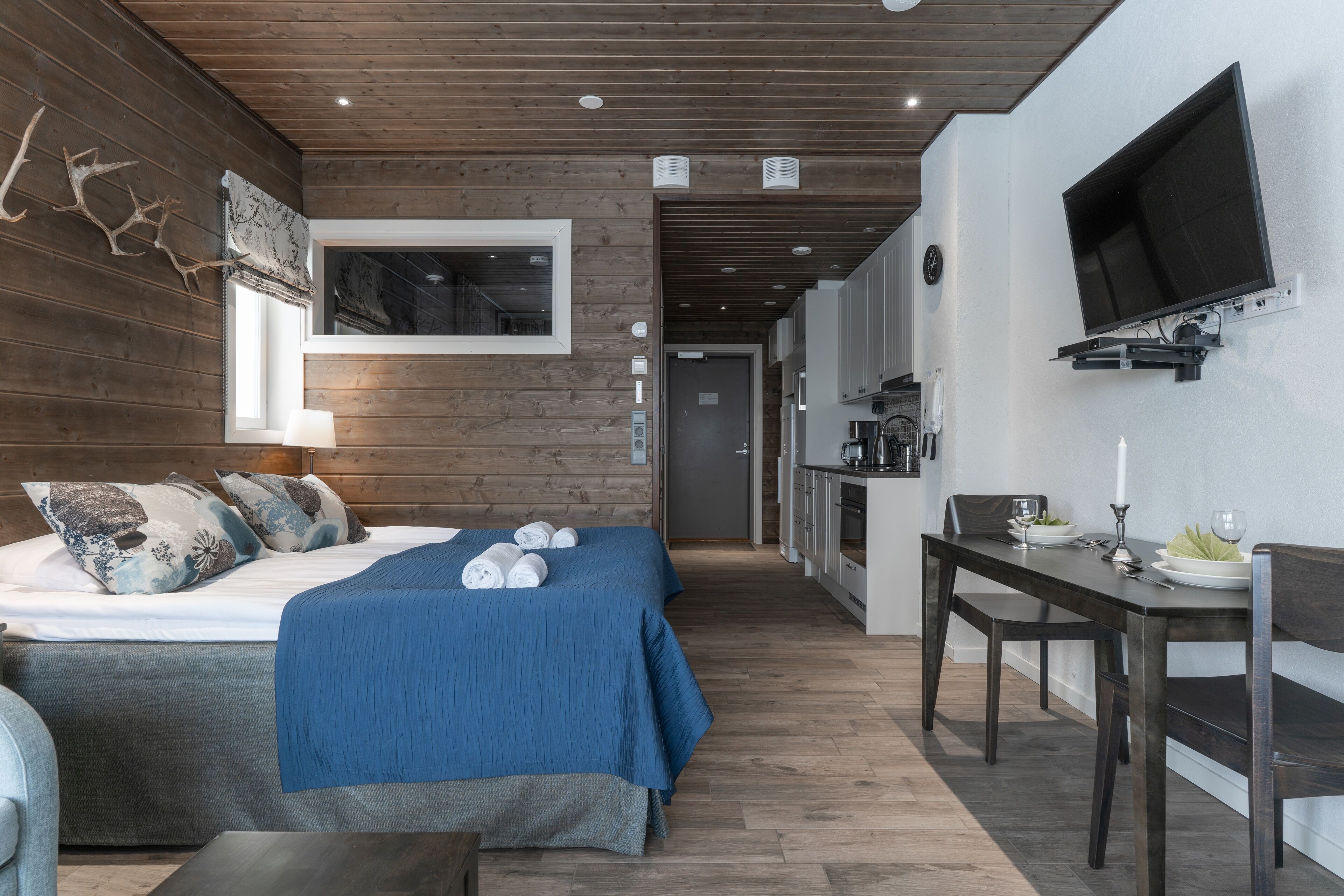 Ein modernes Hotelzimmer mit Holzboden, einem großen Bett mit blauer Bettwäsche, einem Schreibtisch mit Stuhl und einem an der Wand befestigten Fernseher.