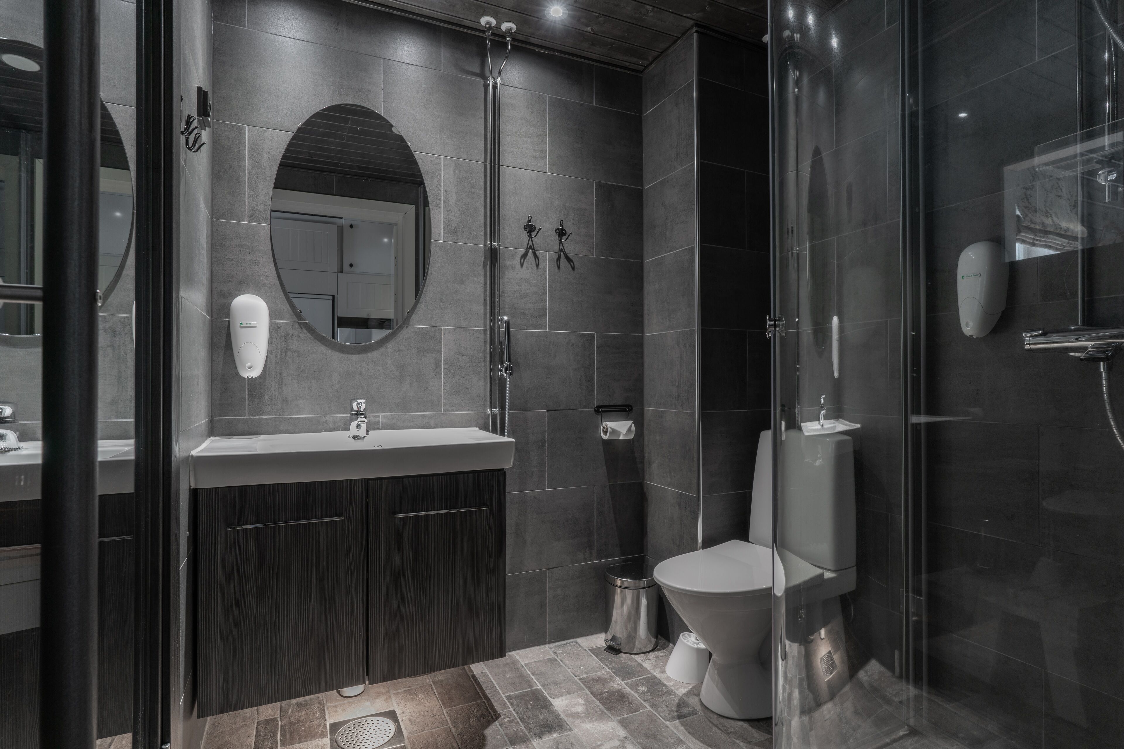 A modern bathroom with a glass shower enclosure, a dark tiled wall, a round mirror, and a toilet with a bidet attachment.