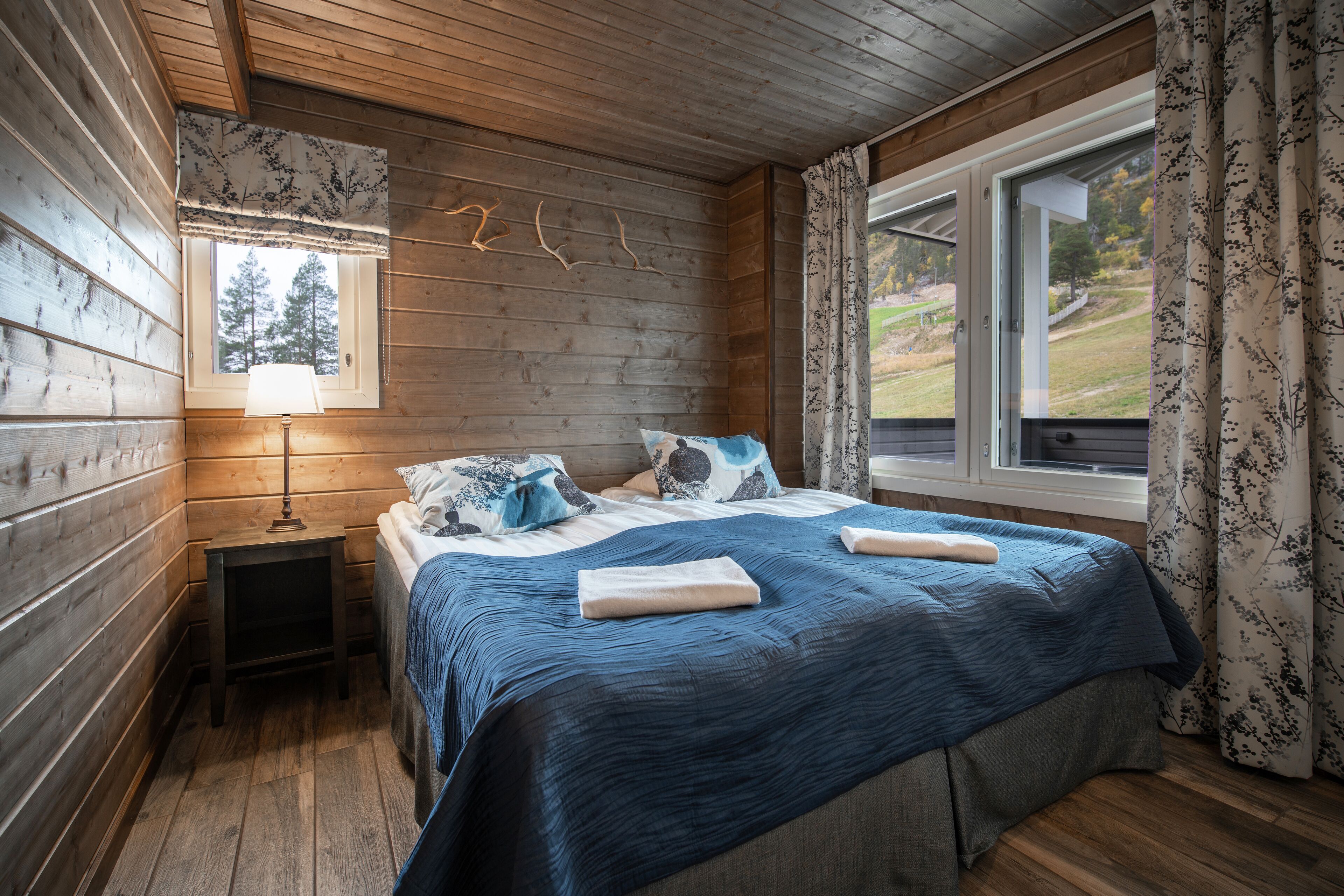 A wooden-paneled room with a bed, a nightstand, a window with curtains, and a view of trees.