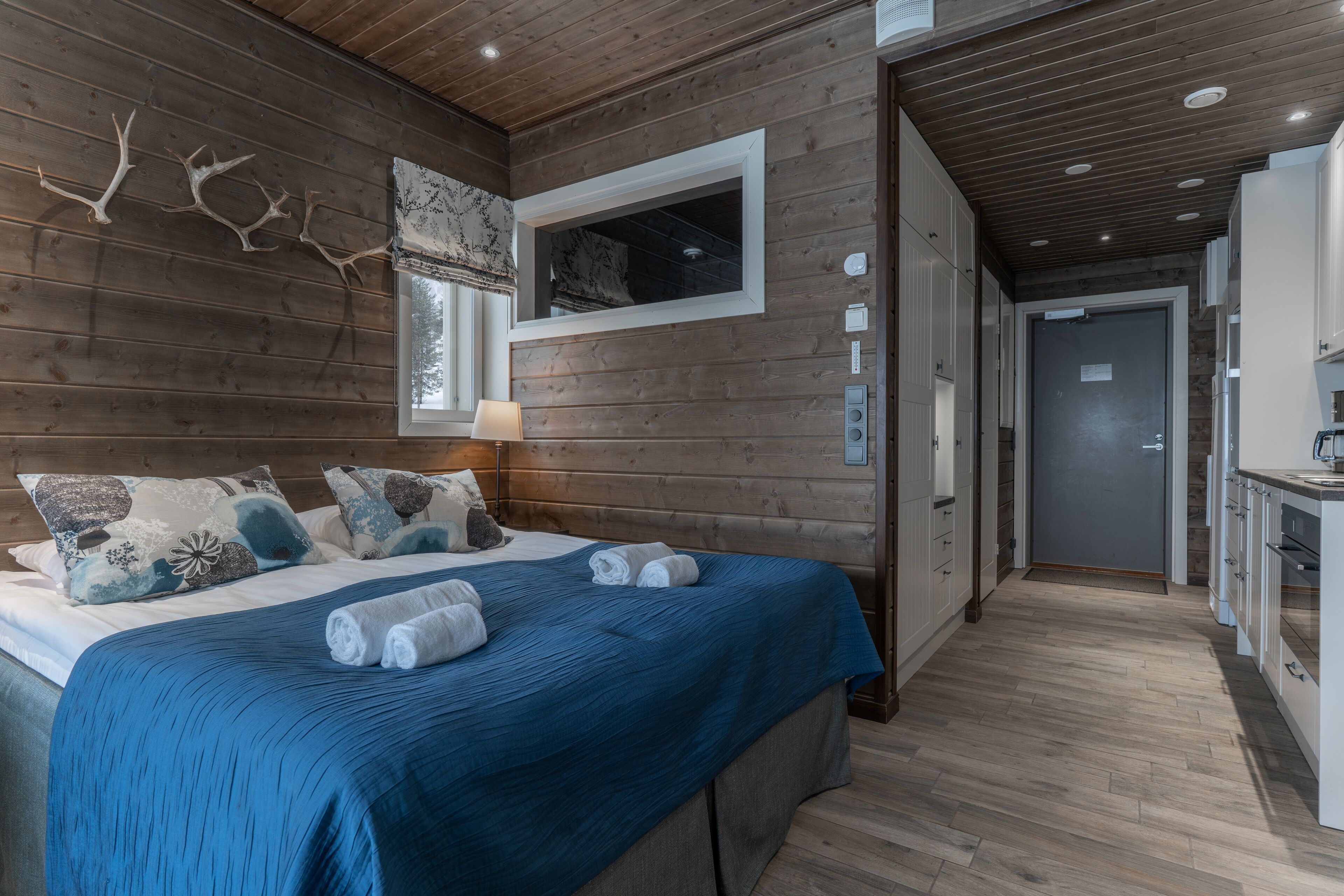Interior de una cabaña de madera con una cama grande, una ventana con vistas y una zona de cocina.