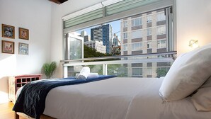 Un dormitorio con una cama grande, vistas de edificios a través de una ventana y una planta en el alféizar.