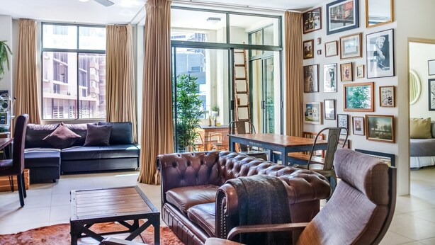A modern living room with a sectional sofa, leather armchair, and a dining area with a wooden table. Large windows offer a view of the cityscape. - ★ Funky Surry Hills Warehouse by ★ Sydney Dreams ★ (Darlinghurst)