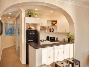 Eine kleine Küche mit weißen Schränken, einem schwarzen Kühlschrank, einem Herd und einem Spülbecken. An der Wand befinden sich Blumentöpfe und eine Weltkarte.