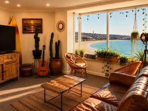 Ein Wohnzimmer mit Blick auf den Strand, einer Gitarre, einem Kaktus und einem Korbstuhl.